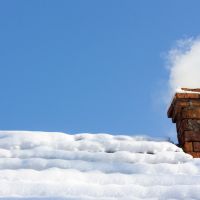 smoke out of a brick chimney on a snowy rooftop on the background of blue sky