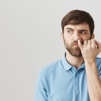 Can I dig to brain. Studio shot of clueless untidy caucasian guy looking aside with confused expression, picking in nose with pinky, thinking nobody sees him over gray background.