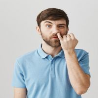 Portrait of funny impolite caucasian man with beard, picking nose with pinky staring with lifted eyebrow at camera, standing over gray background. Wife can not teach husband how to behave in public