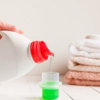 Close up of female hands pouring liquid laundry detergent into cap on white rustic table with towels on background in bathroom.