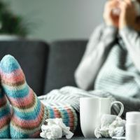 Sick woman with flu, cold, fever and cough sitting on couch at home. Ill person blowing nose and sneezing with tissue and handkerchief. Woolen socks and medicine. Infection in winter. Resting on sofa.