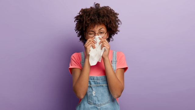 Portrait of sick African American woman sneezes in white tissue, suffers from rhinitis and running nose, has allergy on something, looks unhealthy, feels unwell. Symptoms of cold or allergy.