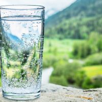 Glass of water on the stone close-up. Blurred snow mountains tops and green forests at the background.