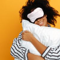 Sleeping. Dreams. Woman portrait. Afro American girl in pajama and sleep mask is hugging a pillow and smiling, on a yellow background