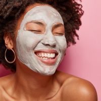Close up portrait of happy delighted woman applies clay mask, closes eyes and smiles delightfully, imagines something pleasant, stands over pink background empty space on right side. Skin care concept